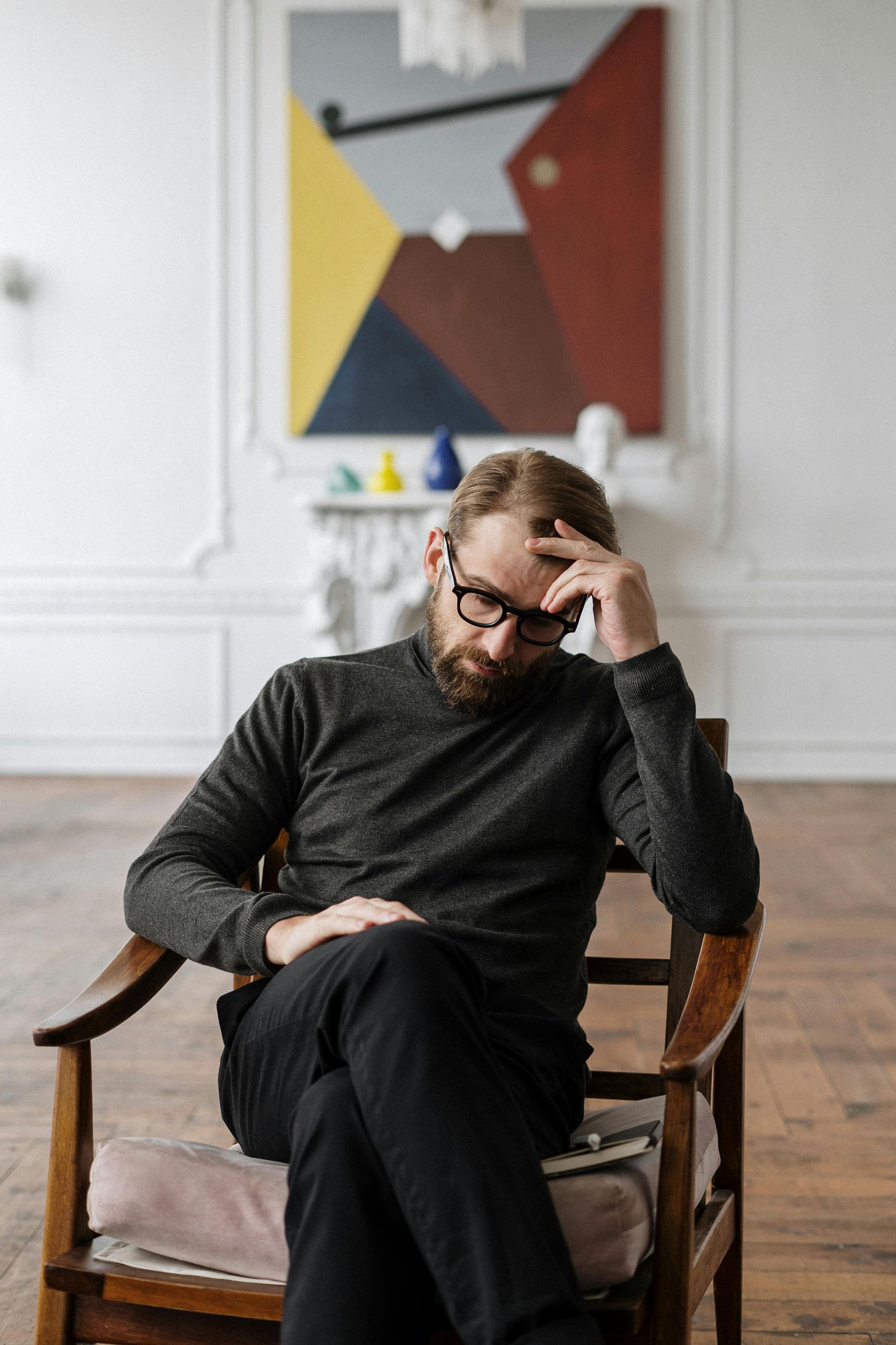 A man with a beard is sitting in a chair in front of a painting.