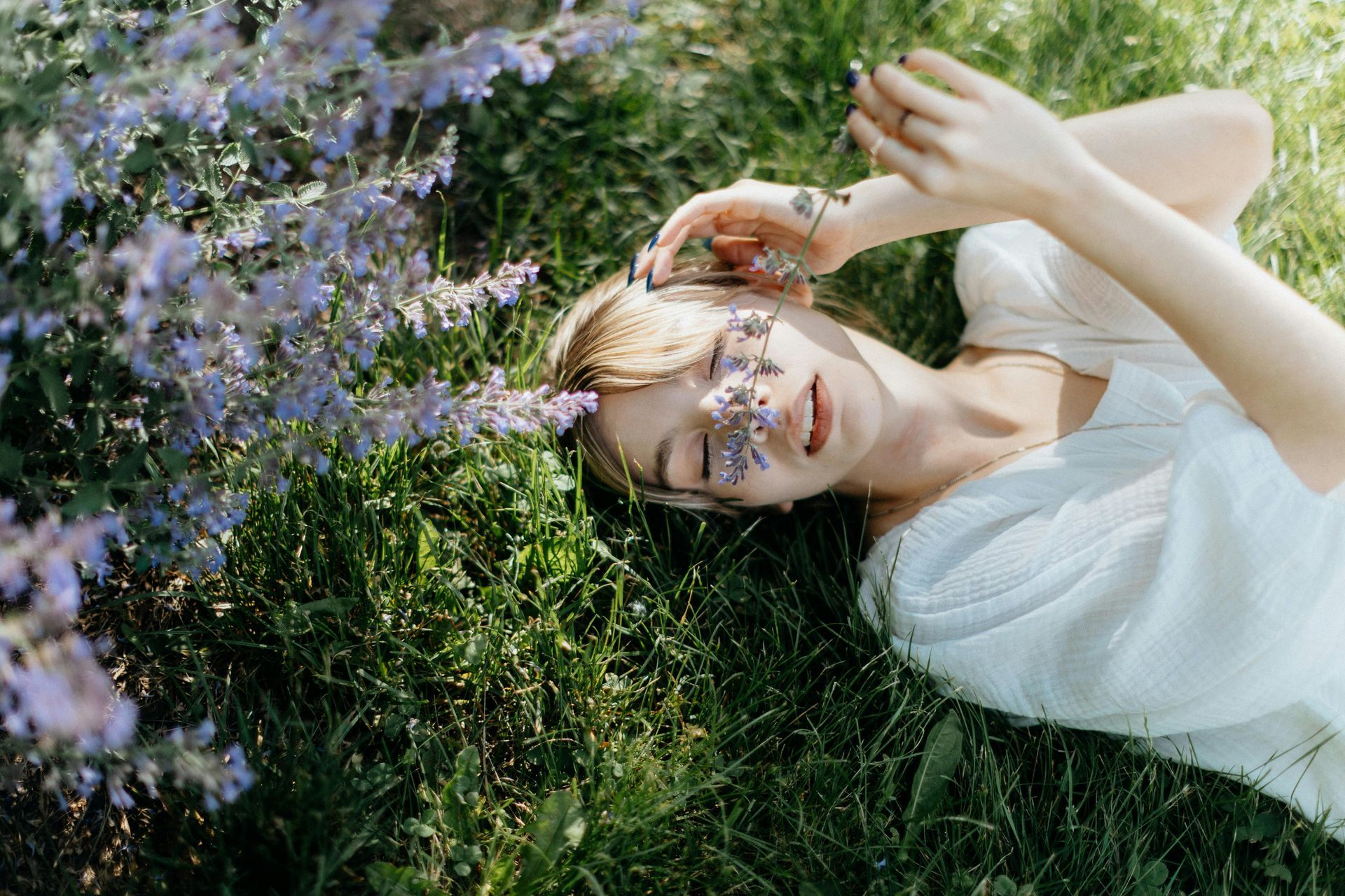 A woman is lying in the grass with purple flowers on her face, smiling and relaxing after a restorative counseling retreat.