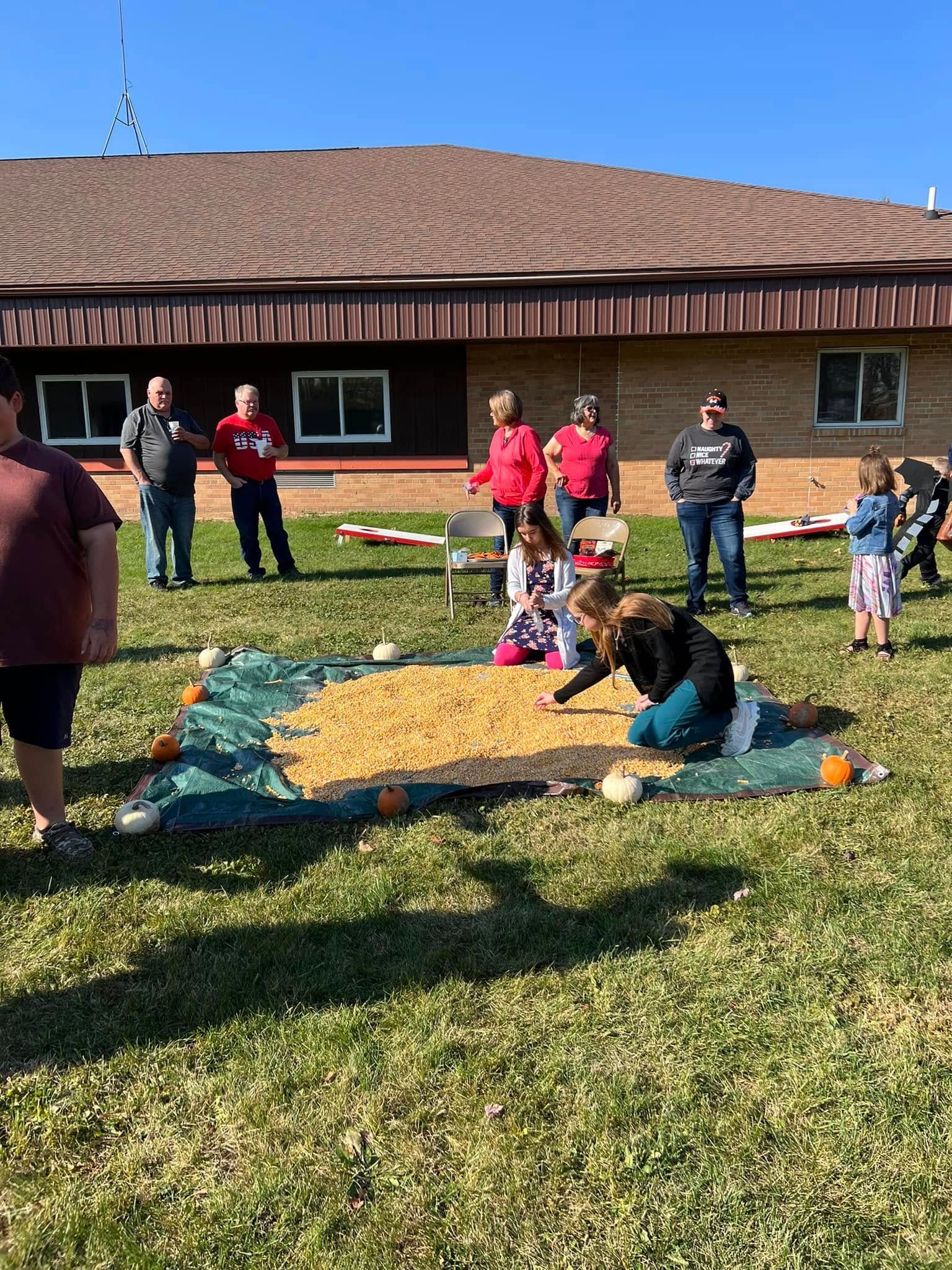 Volunteers sorting donation boxes on a grassy lawn beside a brick building and pickup truck under a blue sky