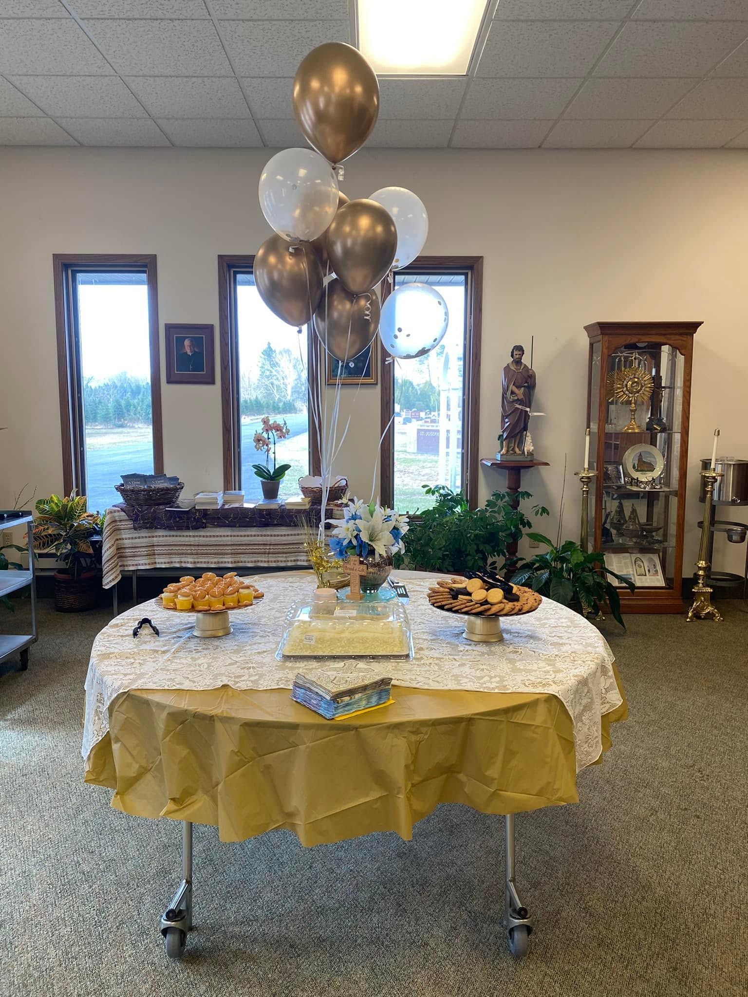 Decorated banquet table with gold balloons, treats, and greenery in a room with display cabinets and windows