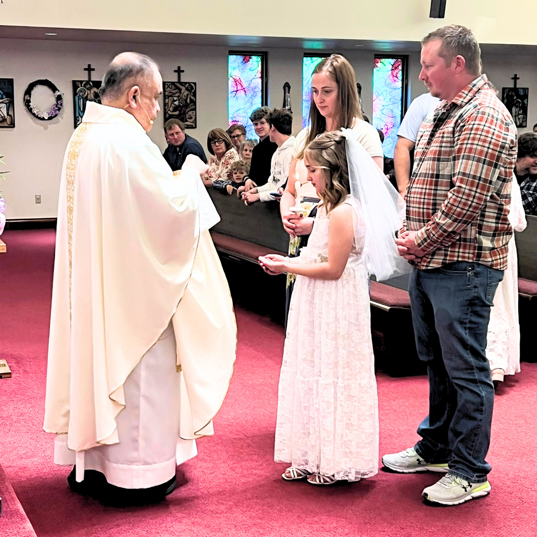 Priest in white robes blessing a young girl in a pink dress at the front of a church sanctuary
