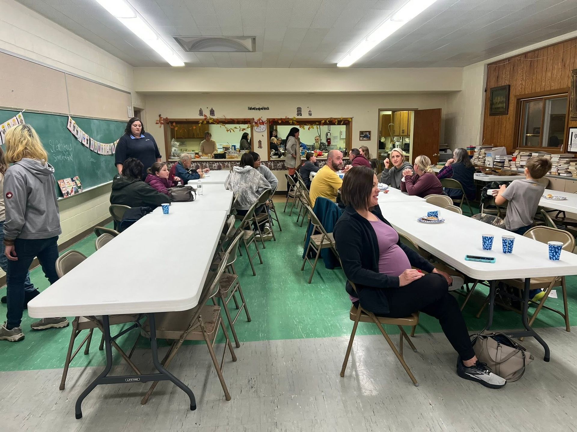 Community gathering in a hall with people seated at long white tables, chatting and eating snacks.