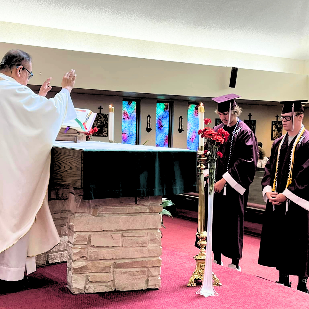 Church ceremony with priest blessing two robed graduates beside an altar