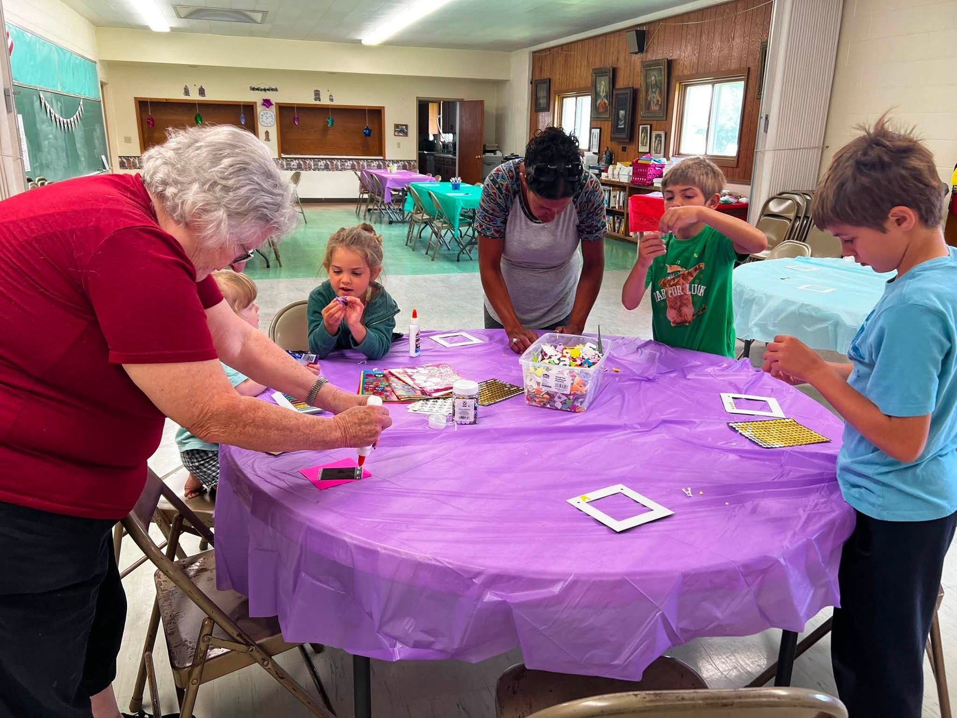 Children and adults make crafts around a purple-covered table in a community room