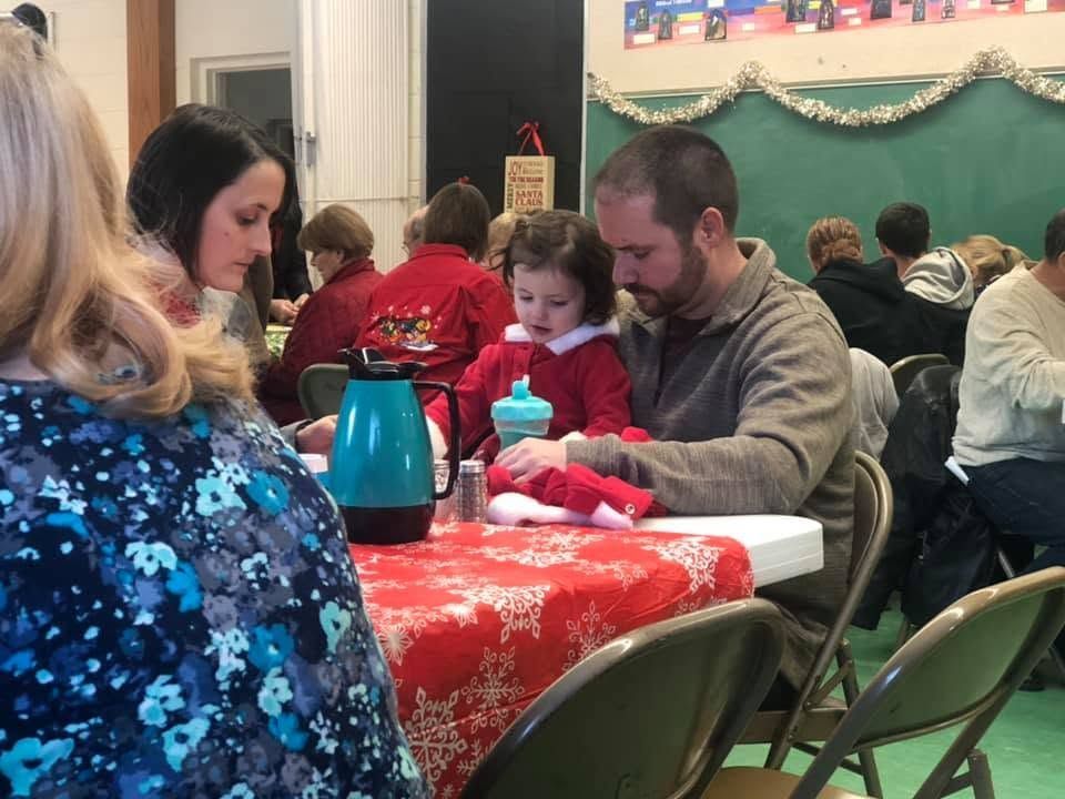 People seated at tables in a decorated hall, with a child and adult sharing a meal at a red tablecloth.