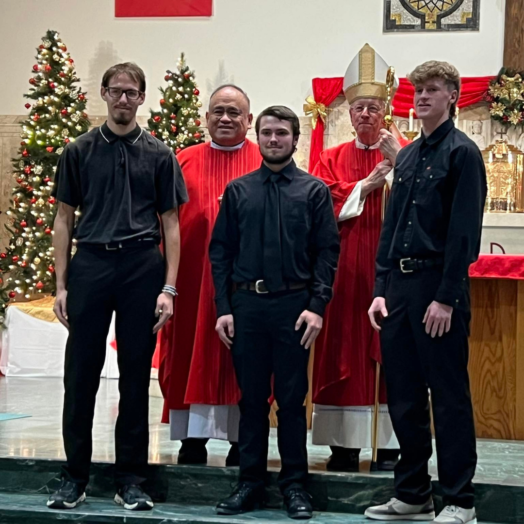 Four men stand in front of a Christmas altar, with decorated trees and clergy in red robes behind them.