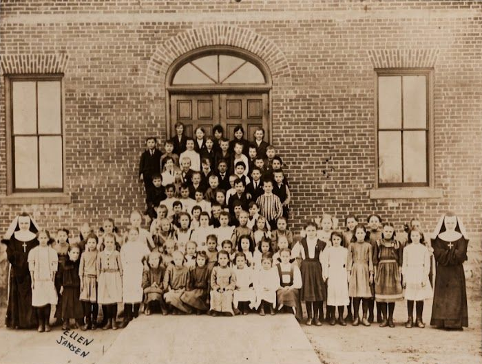 Large group posed on steps outside a brick school building with an arched doorway.