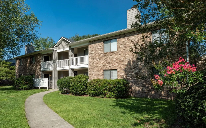 Apartment building with brick facade, white balconies, and a winding path. Green lawn and trees.