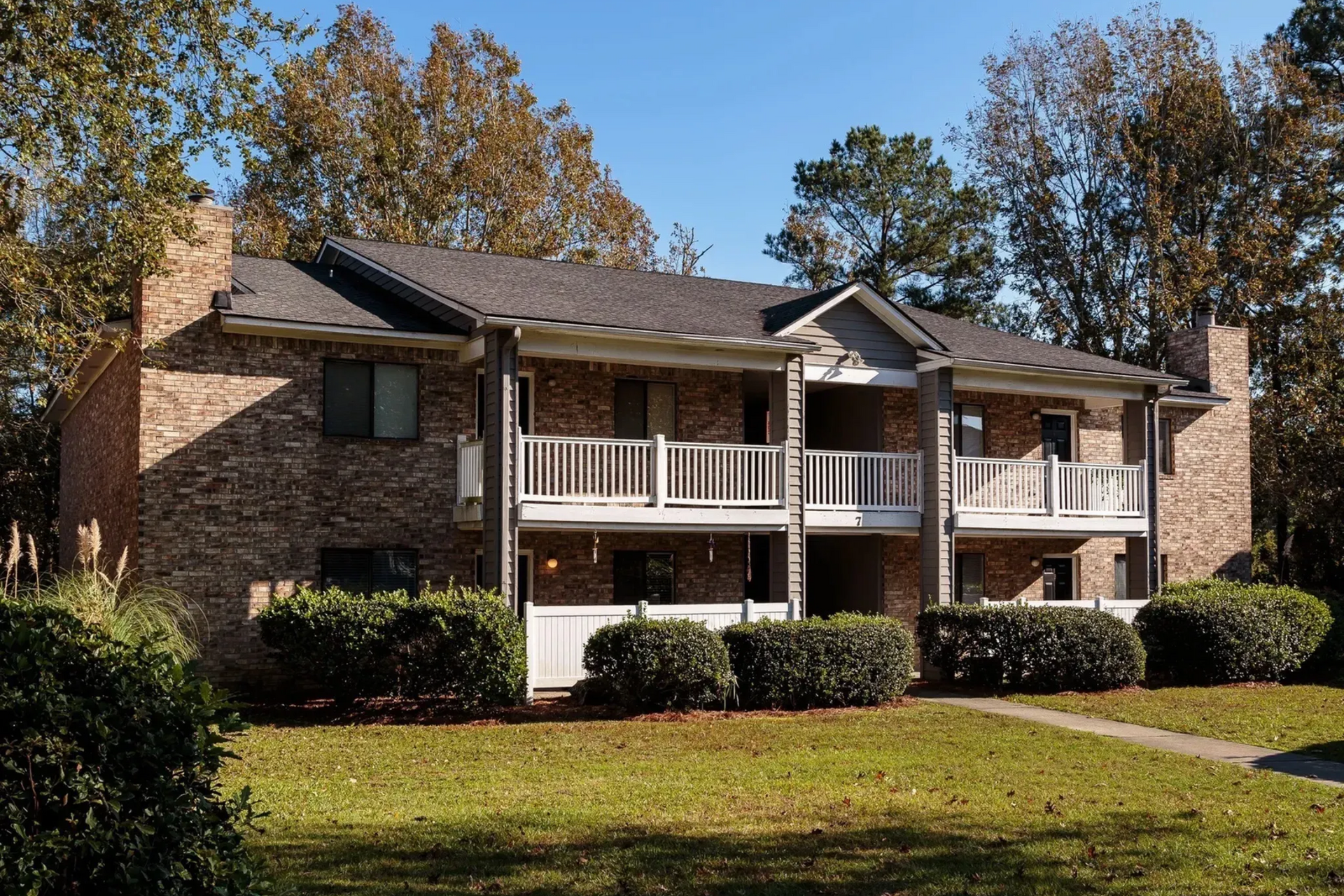Two-story brick apartment building with balconies, surrounded by trees and bushes on a sunny day.