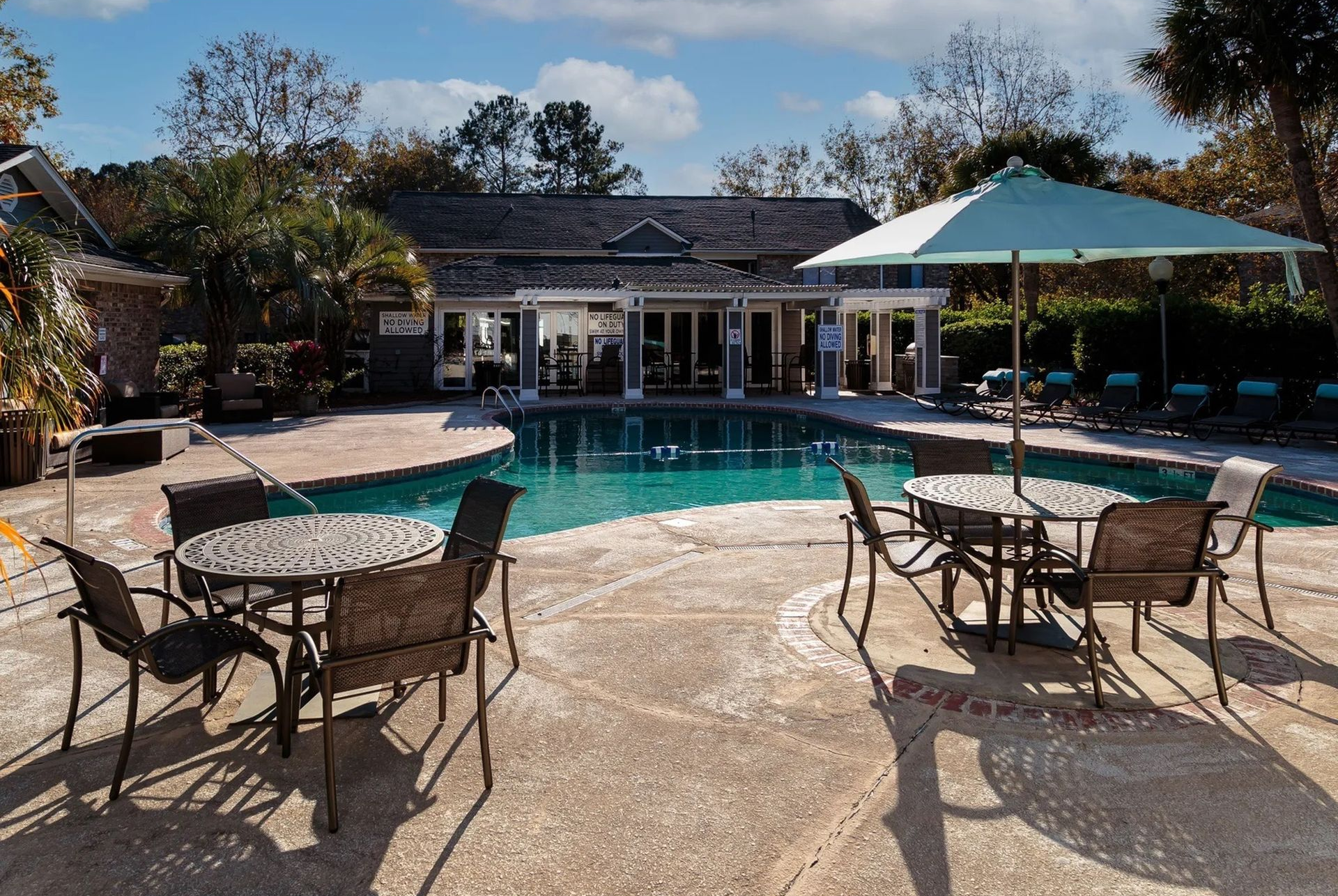 Outdoor community pool area with round metal tables, chairs, and a large umbrella.