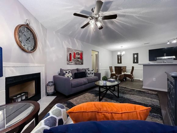 Open-concept living room in an apartment with a sofa, fireplace, large wall clock, ceiling fan, and dining area.