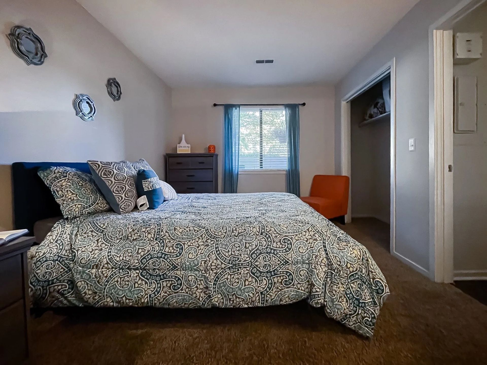 Bedroom in an apartment with a bed, dresser, window with blue curtains, and an open closet.