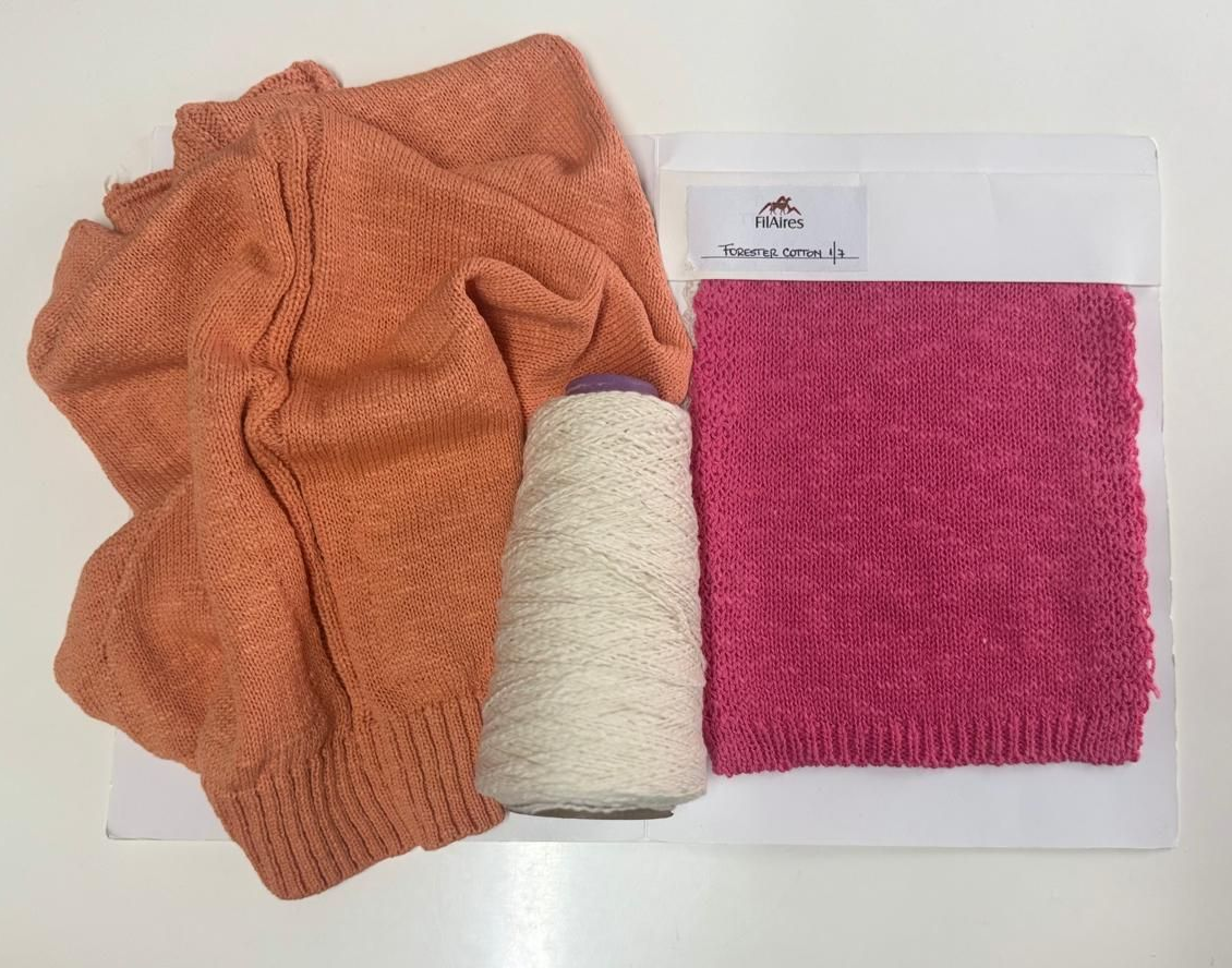 Suéter naranja, cono de hilo blanco y una muestra de punto rosa sobre una superficie blanca.