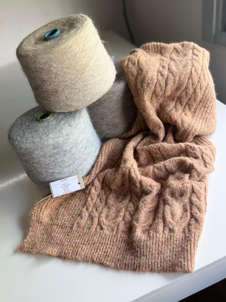 Carretes de hilo gris, beige y tostado junto a una bufanda de punto color melocotón con diseño de trenzas.imagen