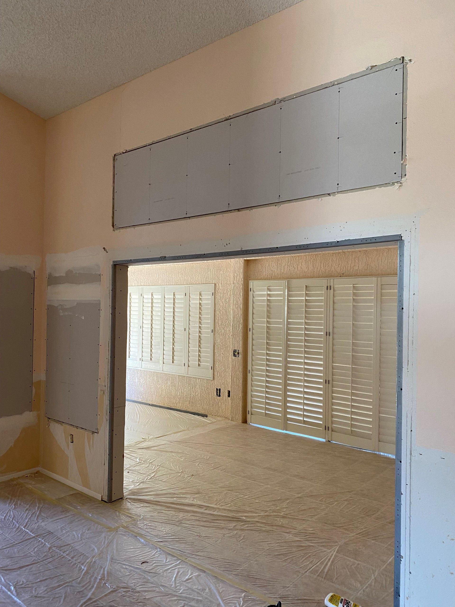 Interior view of a room under renovation. Drywall is installed around doorway and ceiling, with a view of shuttered windows.