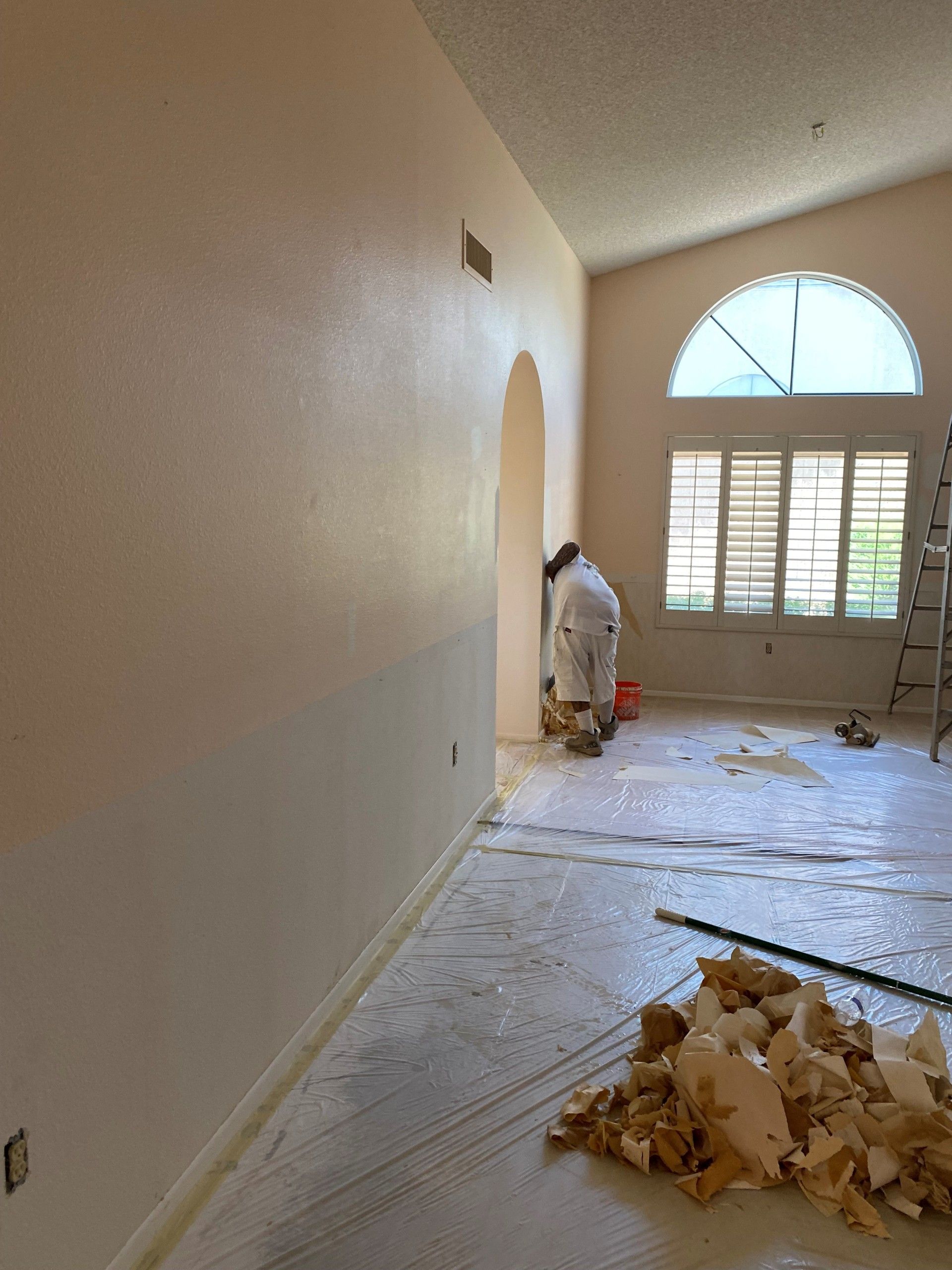 Person painting interior wall; light tan walls, white protective covering on floor, arched doorway, window.