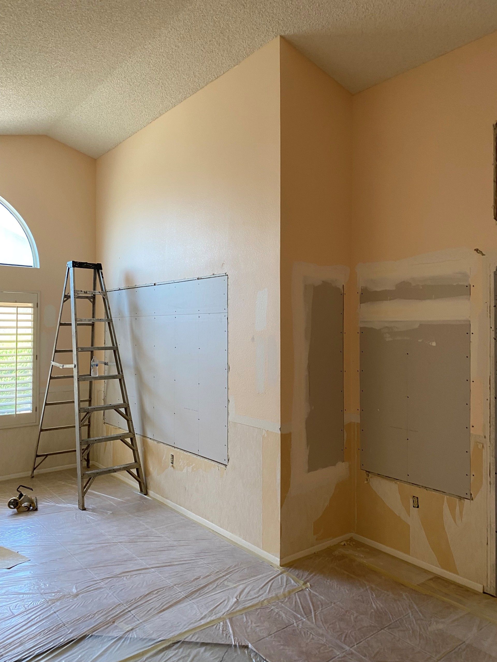 Interior room undergoing renovation; peach walls, ladder, covered floor. Drywall on walls, window with blinds.