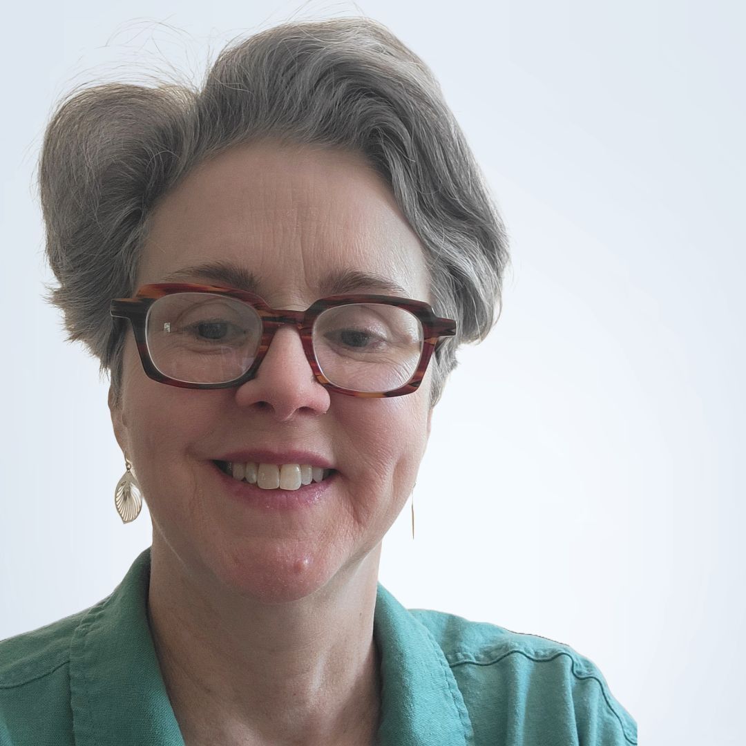 Woman with gray hair and glasses smiles, wearing a green shirt, looking at the camera.