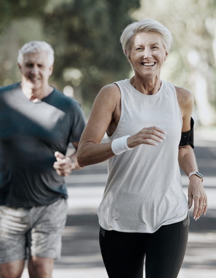 Couple jogging outdoors, woman smiling, wearing a white tank top and black leggings, man in a blue shirt.