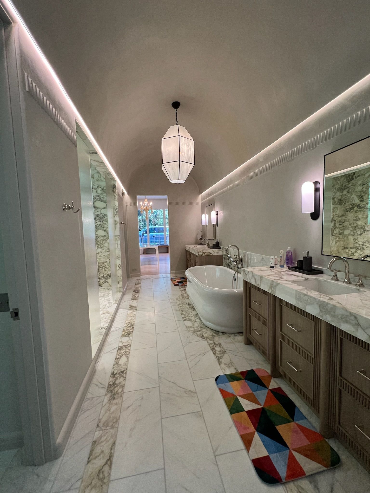 Long, bright bathroom with marble floors, freestanding tub, vanity, colorful rug, and pendant light.