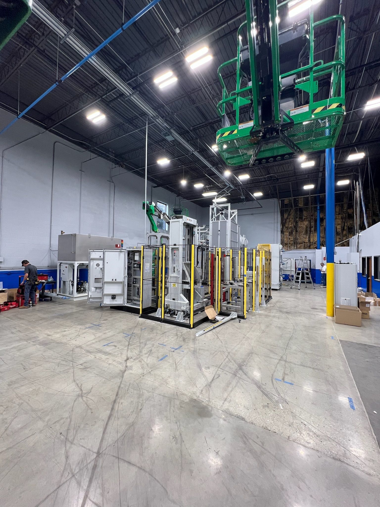 Warehouse interior with tall storage racks, green machinery overhead, and a worker near equipment.