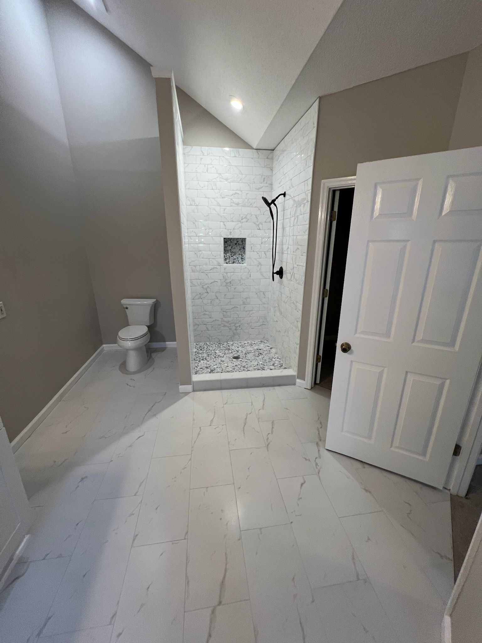 Modern bathroom with white tile, toilet, walk-in shower, and open door near vanity area