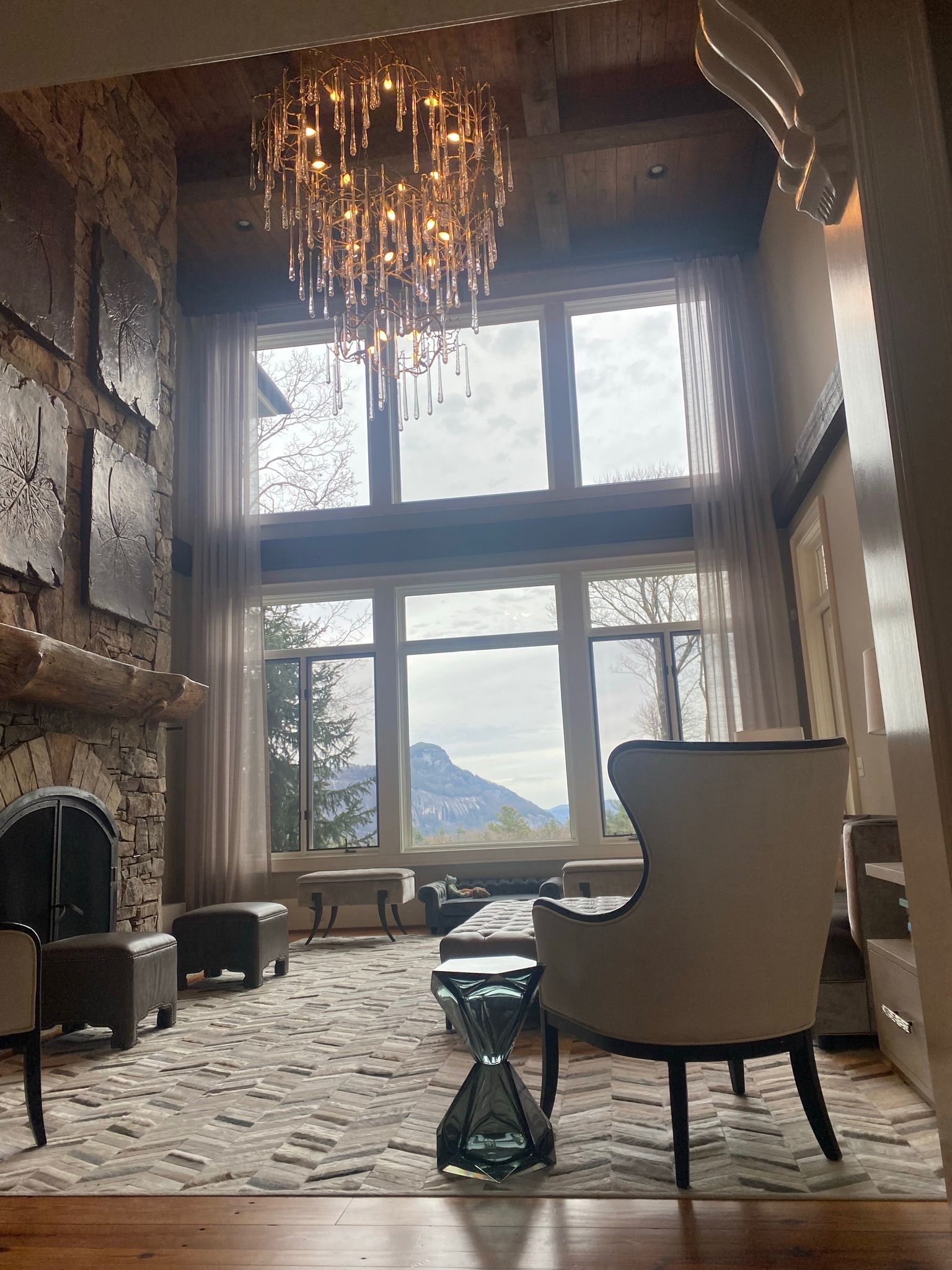 Elegant chalet living room with tall windows, chandelier, stone fireplace, and mountain view