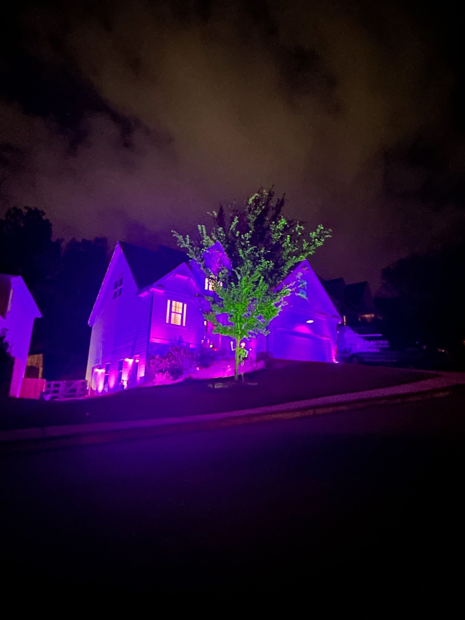 House and trees lit in vivid purple and pink at night under a cloudy sky