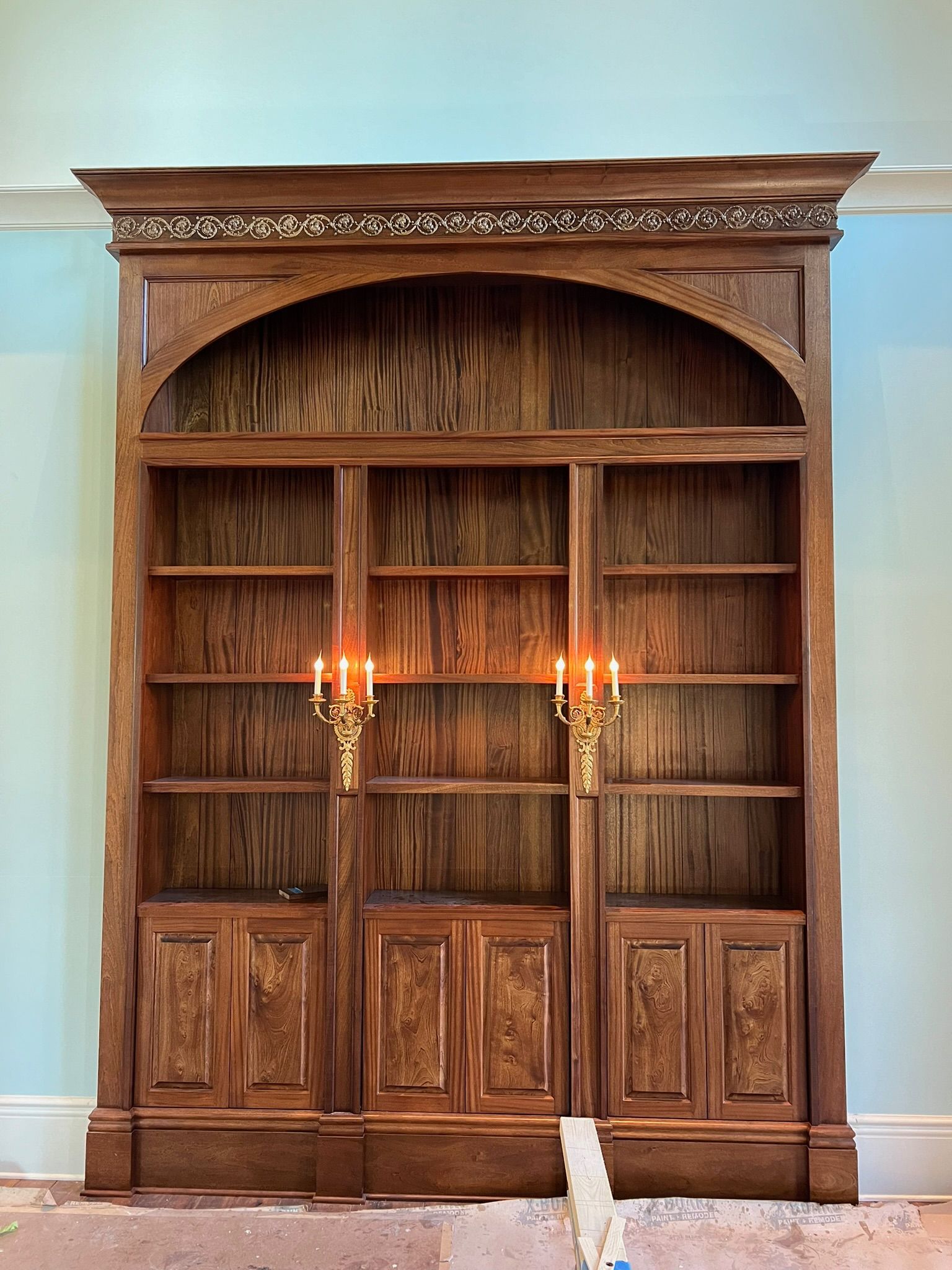 Tall wooden built-in bookshelf with two lit wall sconces and an arched top niche