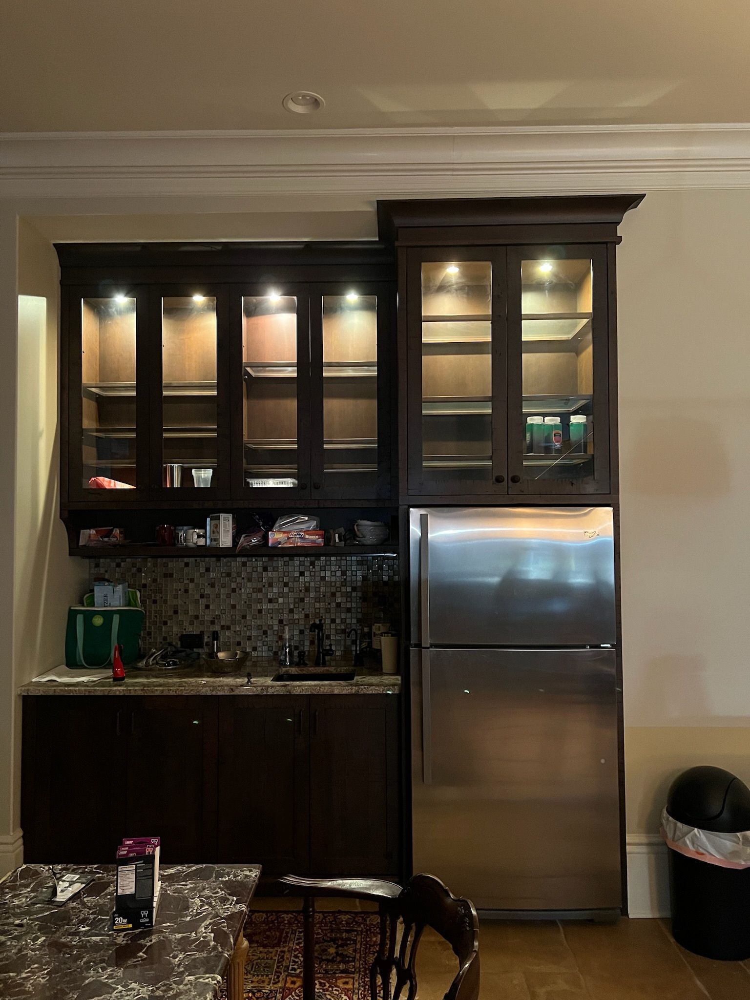 Modern kitchen with dark cabinets, under-cabinet lights, and a stainless-steel refrigerator; dining table in foreground
