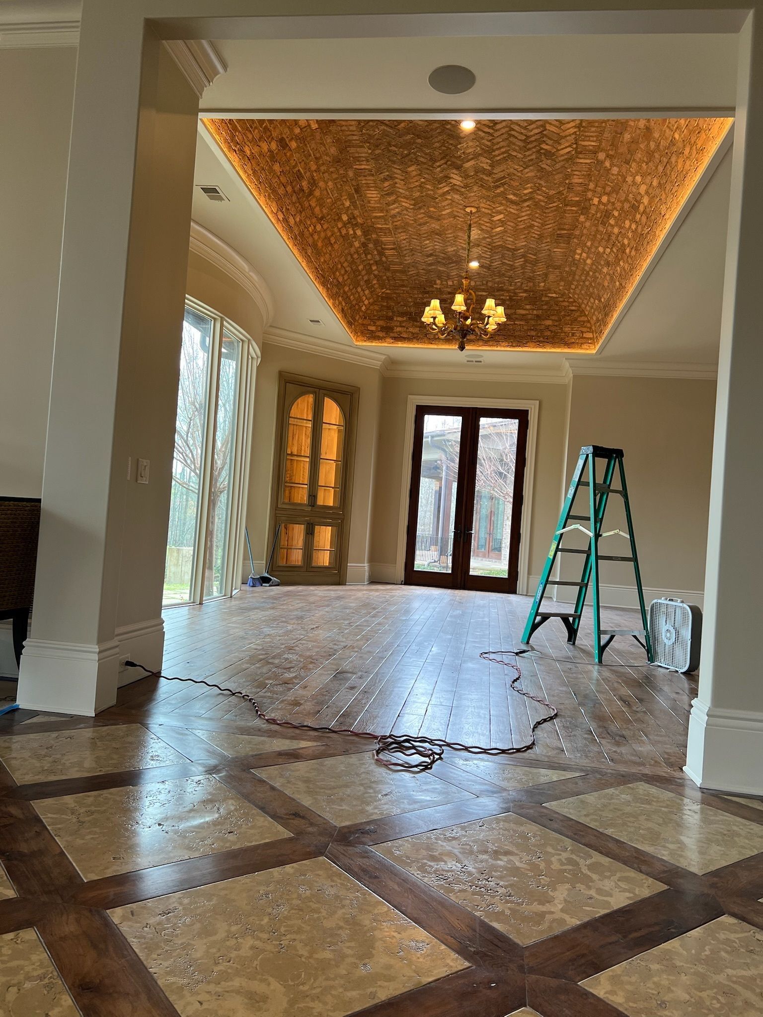 Empty elegant foyer with patterned marble floor, ornate ceiling, chandelier, and a ladder near the back doors