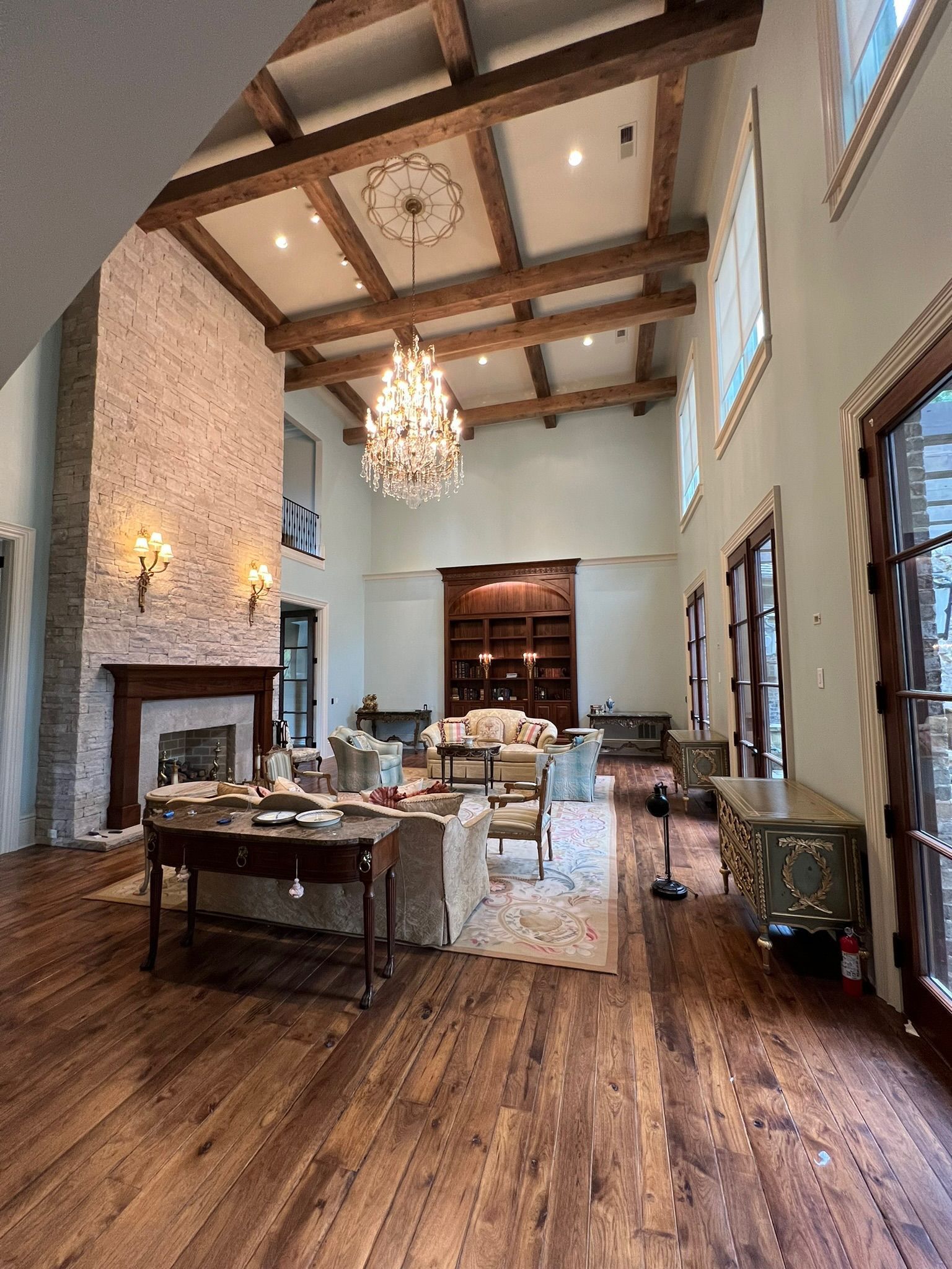 Grand living room with wood floors, exposed beams, chandelier, fireplace, and large windows