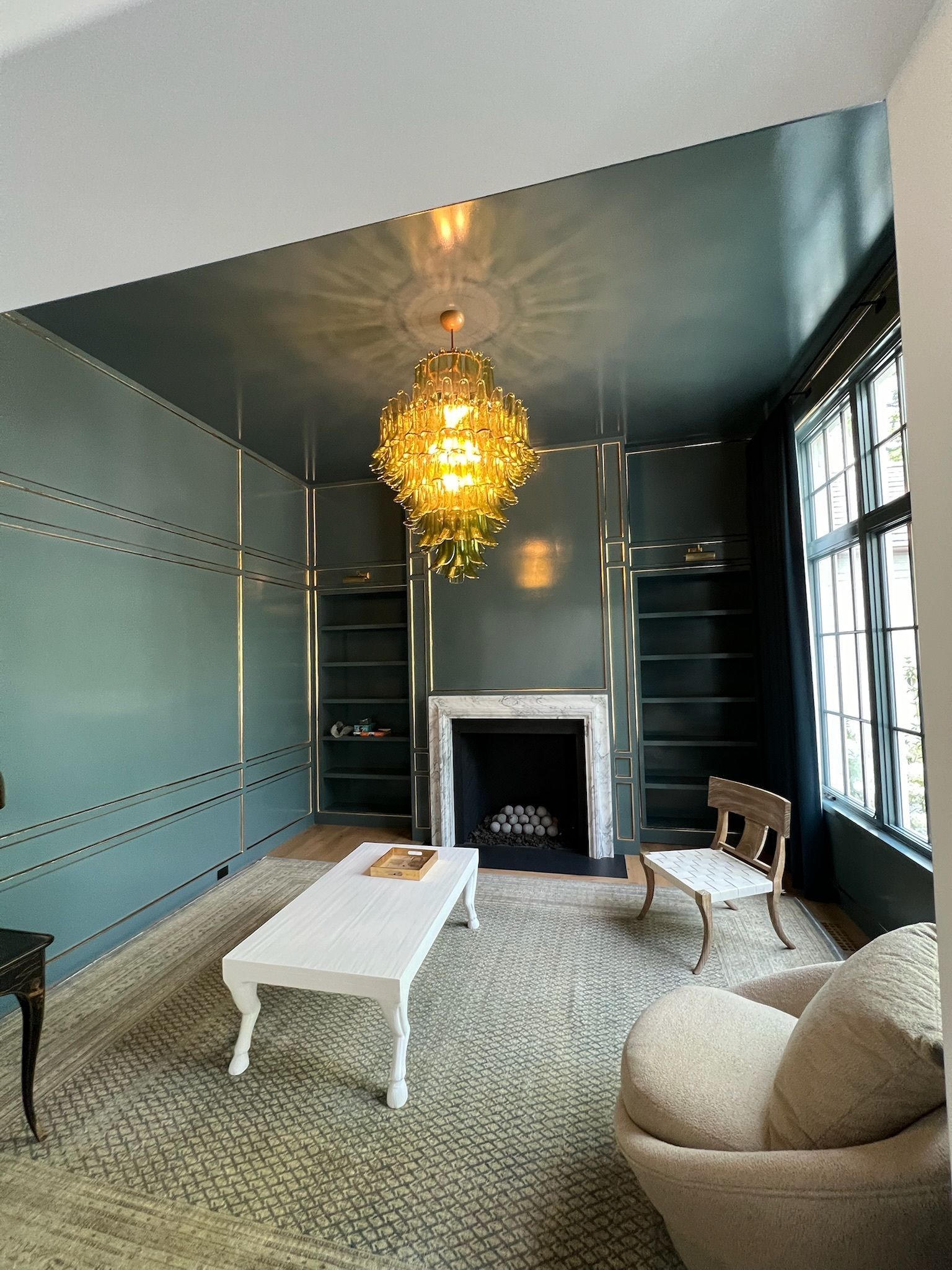 Modern living room with teal walls, gold chandelier, fireplace, white coffee table, and cream armchairs