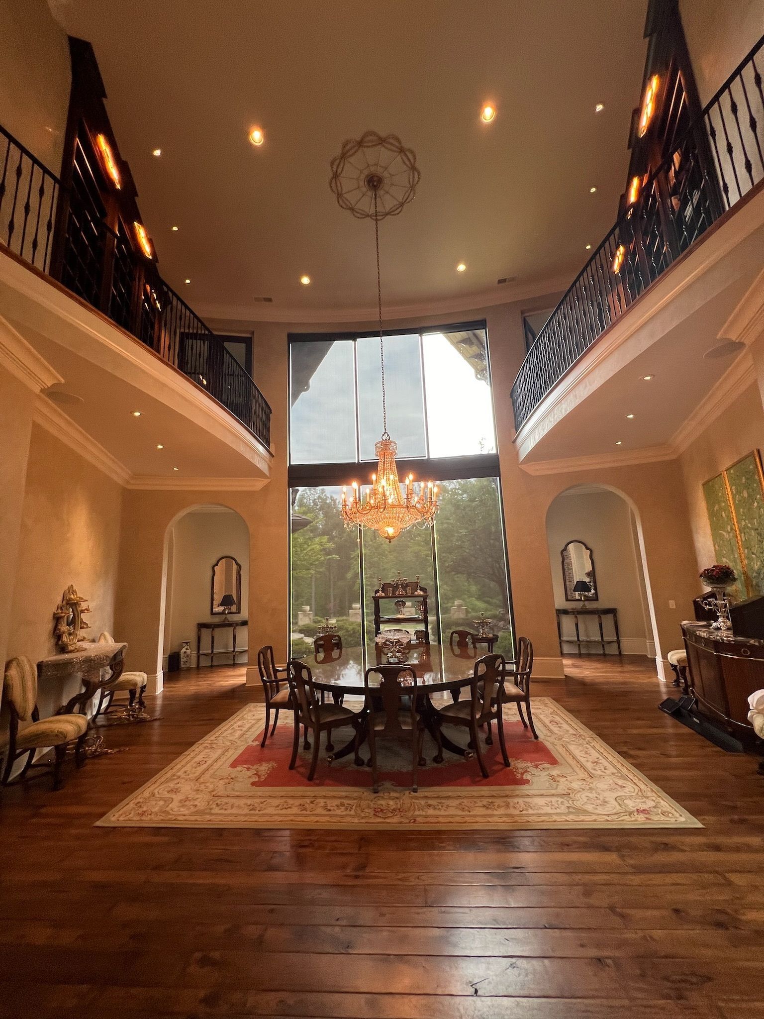 Grand two-story living room with tall windows, chandelier, warm wood floors, and a central dining table