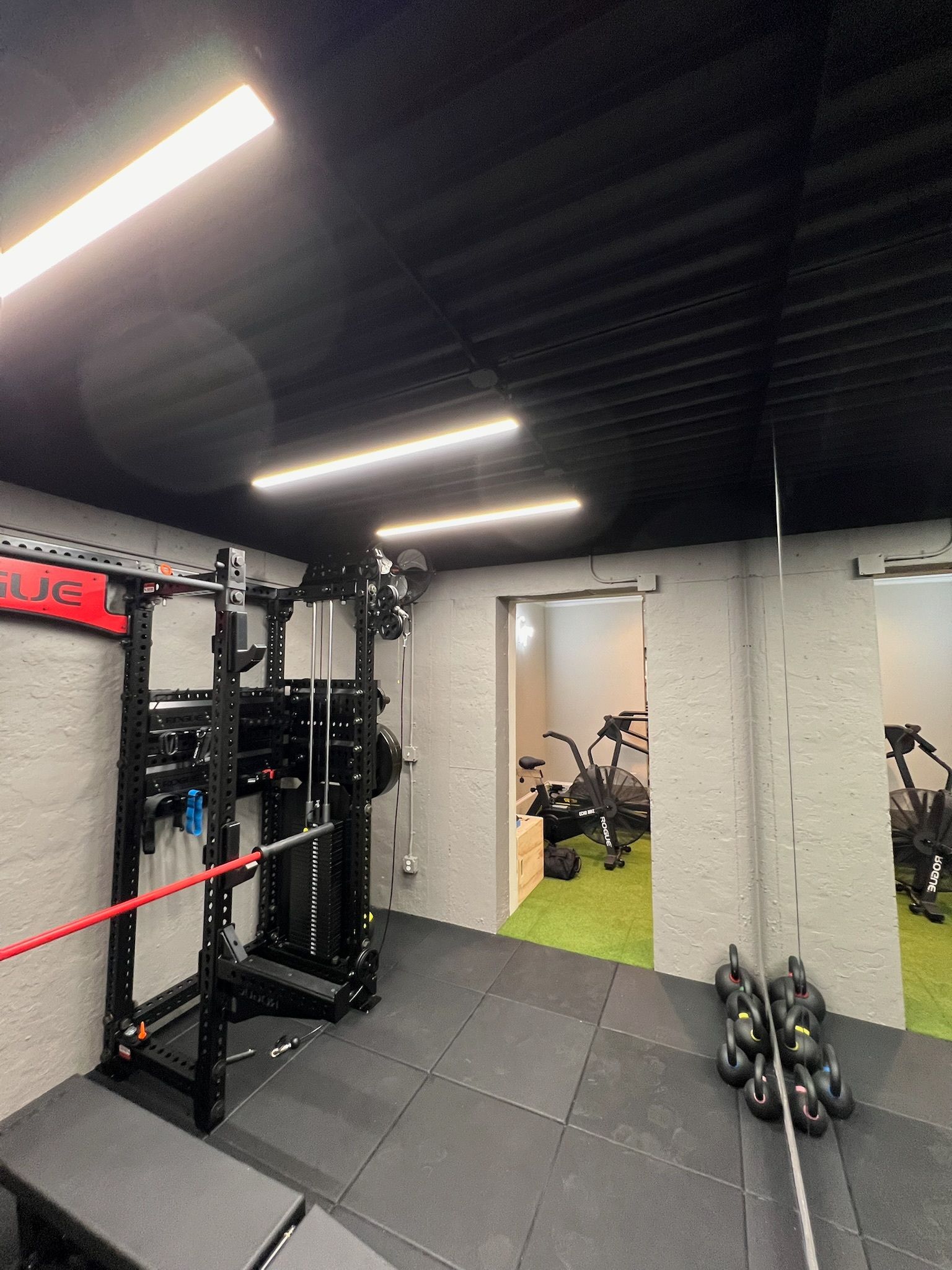 Small gym interior with weight equipment, mats, dumbbells, and an open doorway to a brighter room