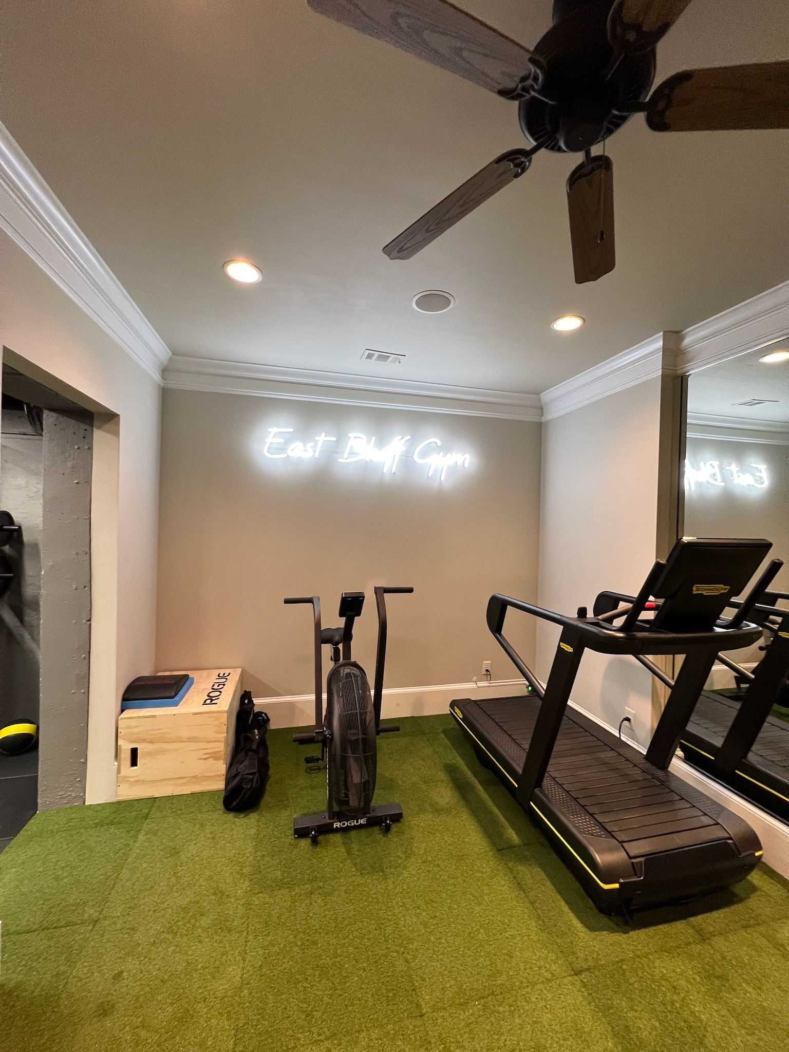 Home gym with treadmill, stationary bike, and green turf flooring under a ceiling fan and wall sign.