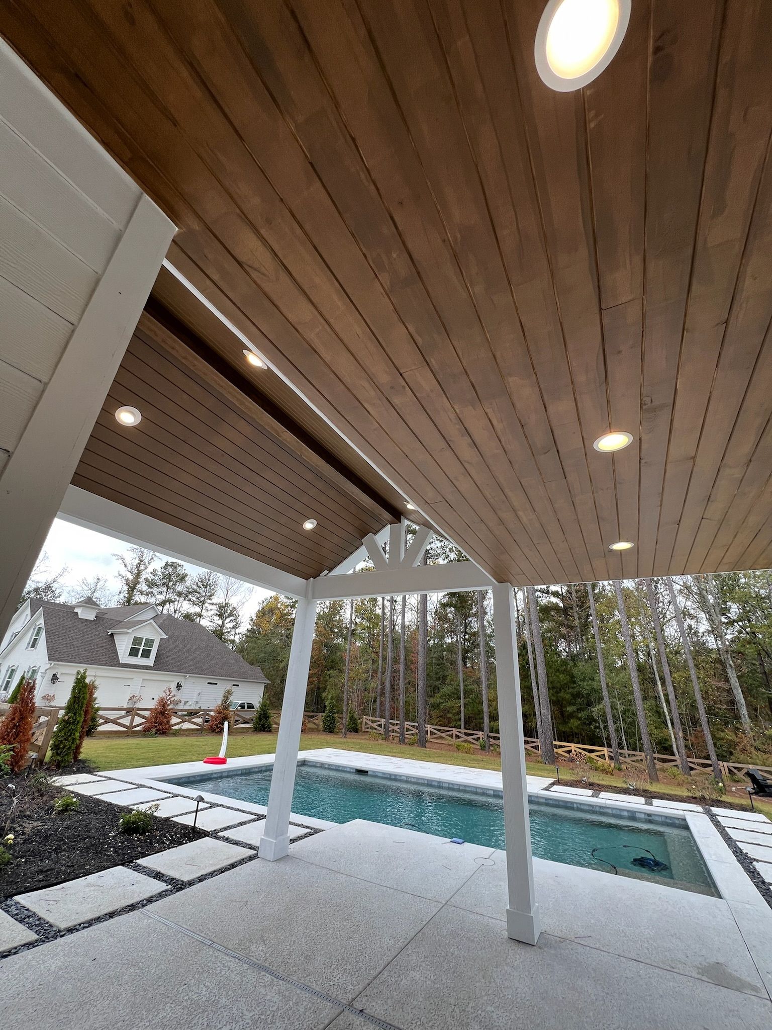 Covered patio beside a backyard pool, with white columns, wood ceiling, and trees beyond