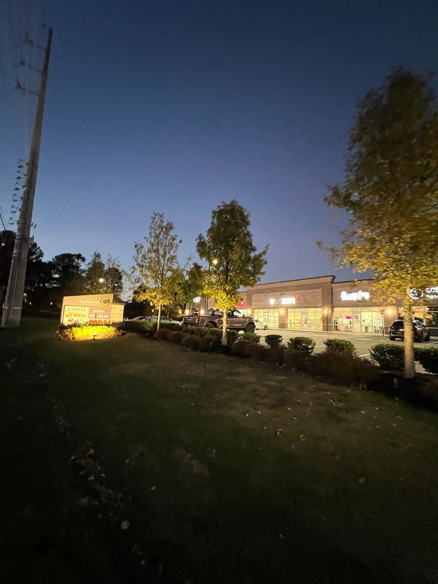Dusk view of a grassy roadside with lit houses, trees, and a yellow sign by the street.