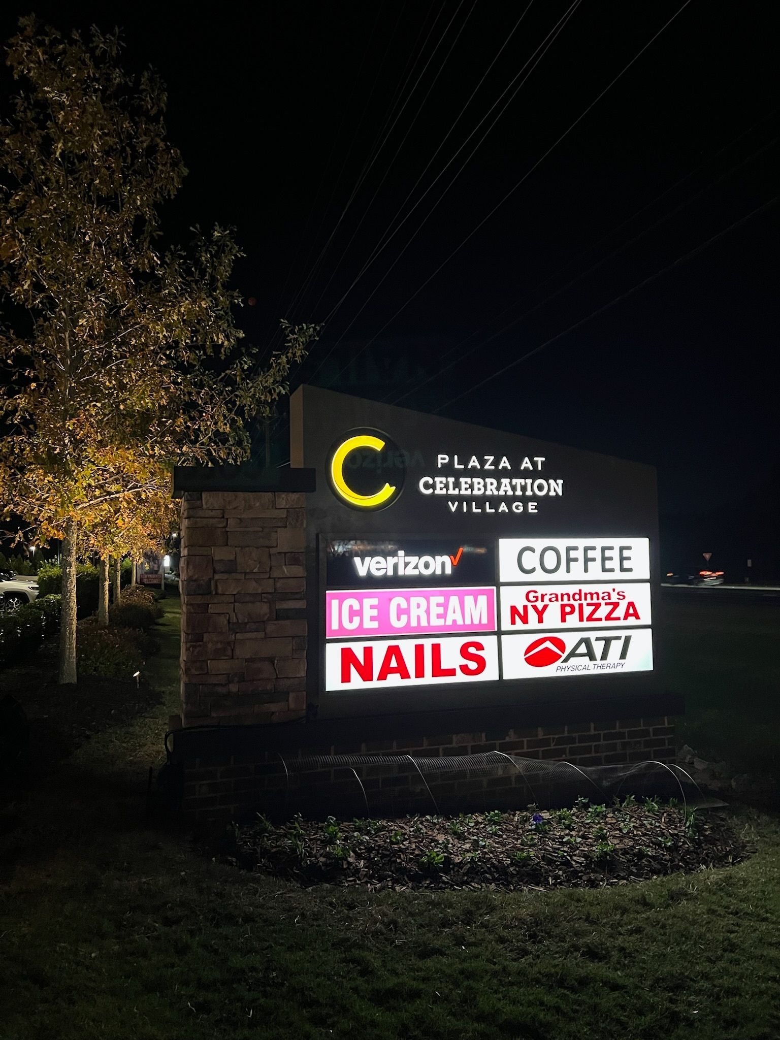 Outdoor plaza sign at night listing Verizon, ice cream, nails, coffee, and pizza businesses