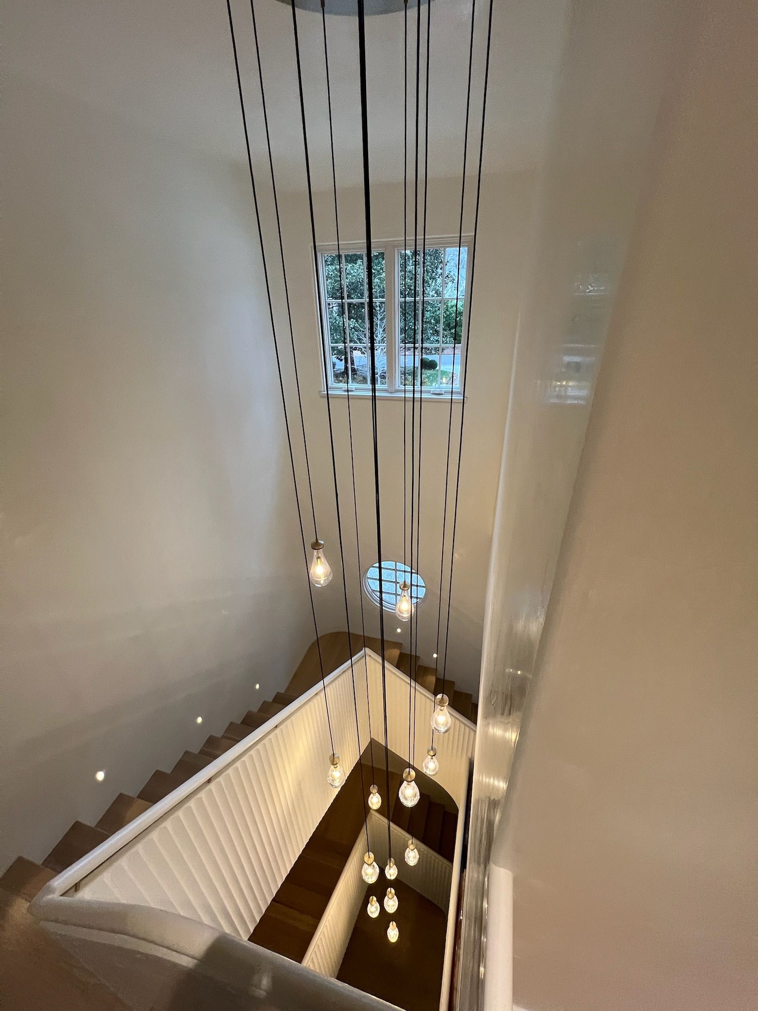 Tall stairwell with hanging lights, white walls, and a window at the top and bottom.