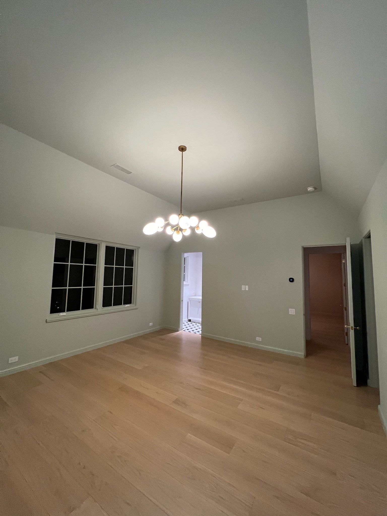 Empty living room with wood floor, white walls, chandelier, windows, and open doorway to hall