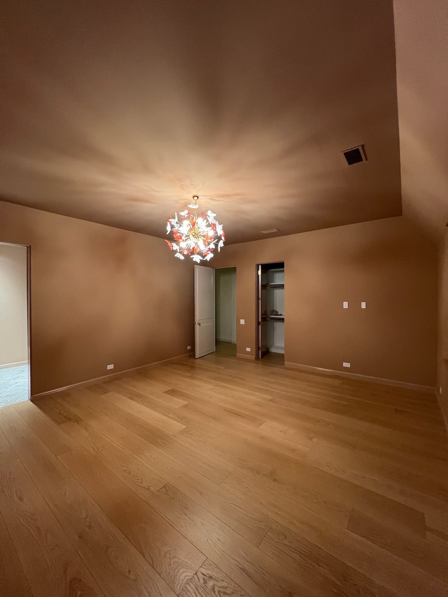 Empty tan-walled room with wood flooring and a hanging chandelier, viewed toward an open doorway.
