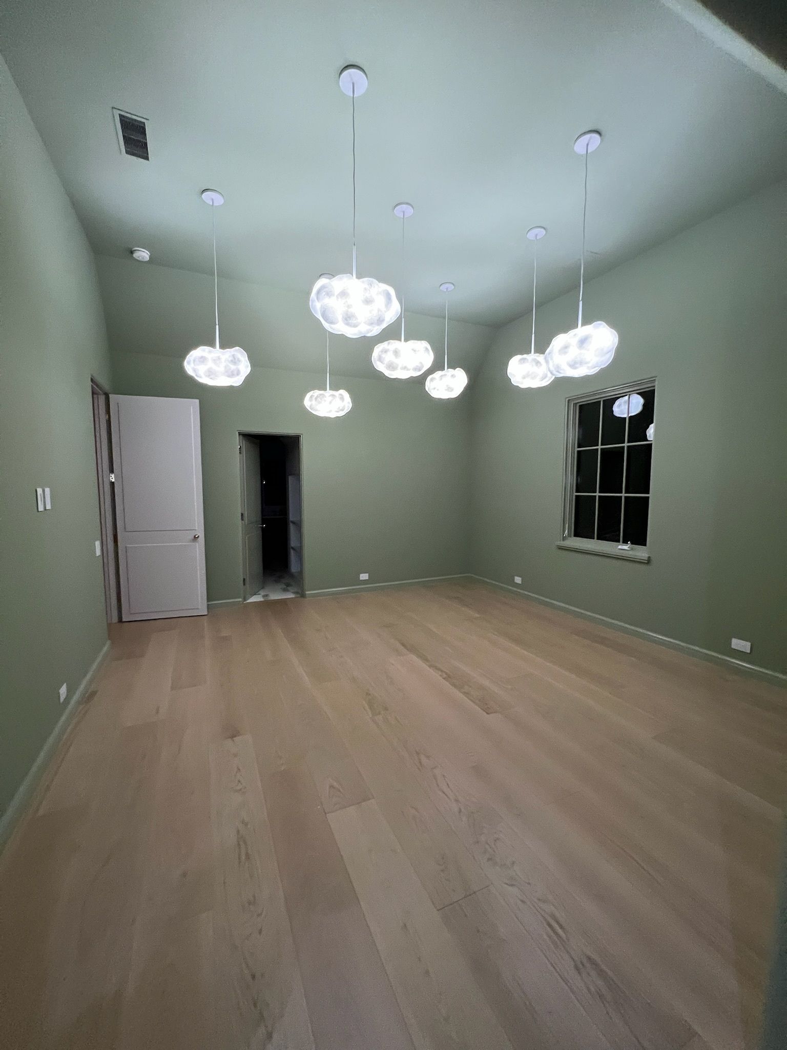 Empty modern room with pale green walls, wood floors, hanging pendant lights, and a window on the right.