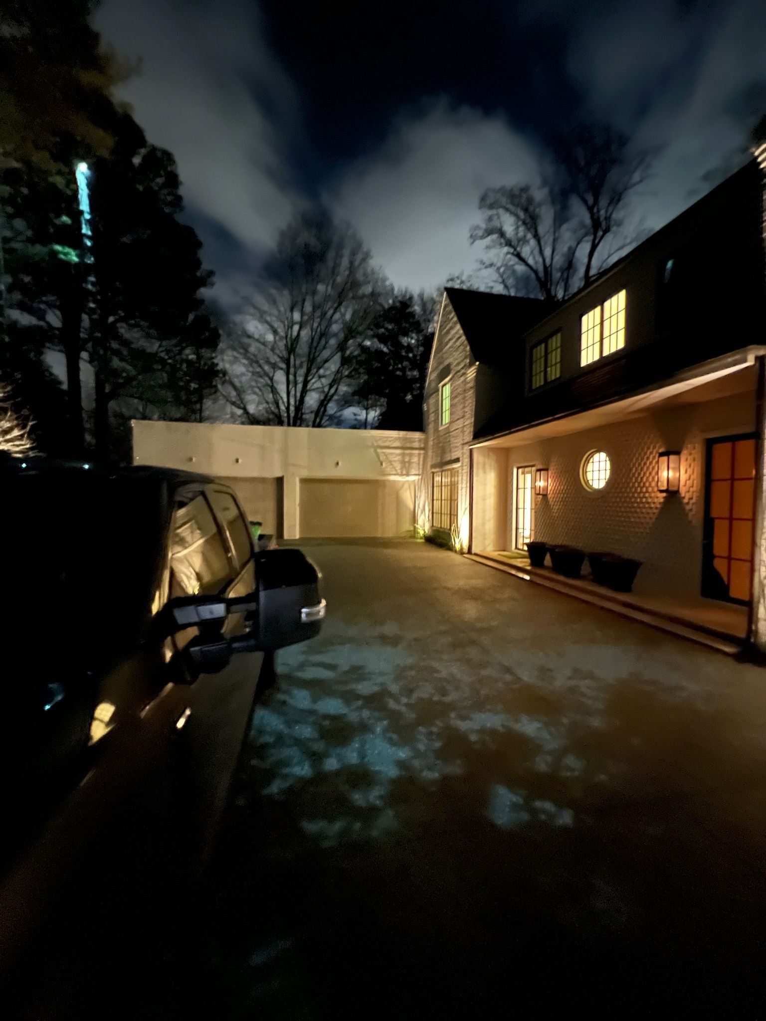 Nighttime driveway beside a lit brick house and parked car under a cloudy sky