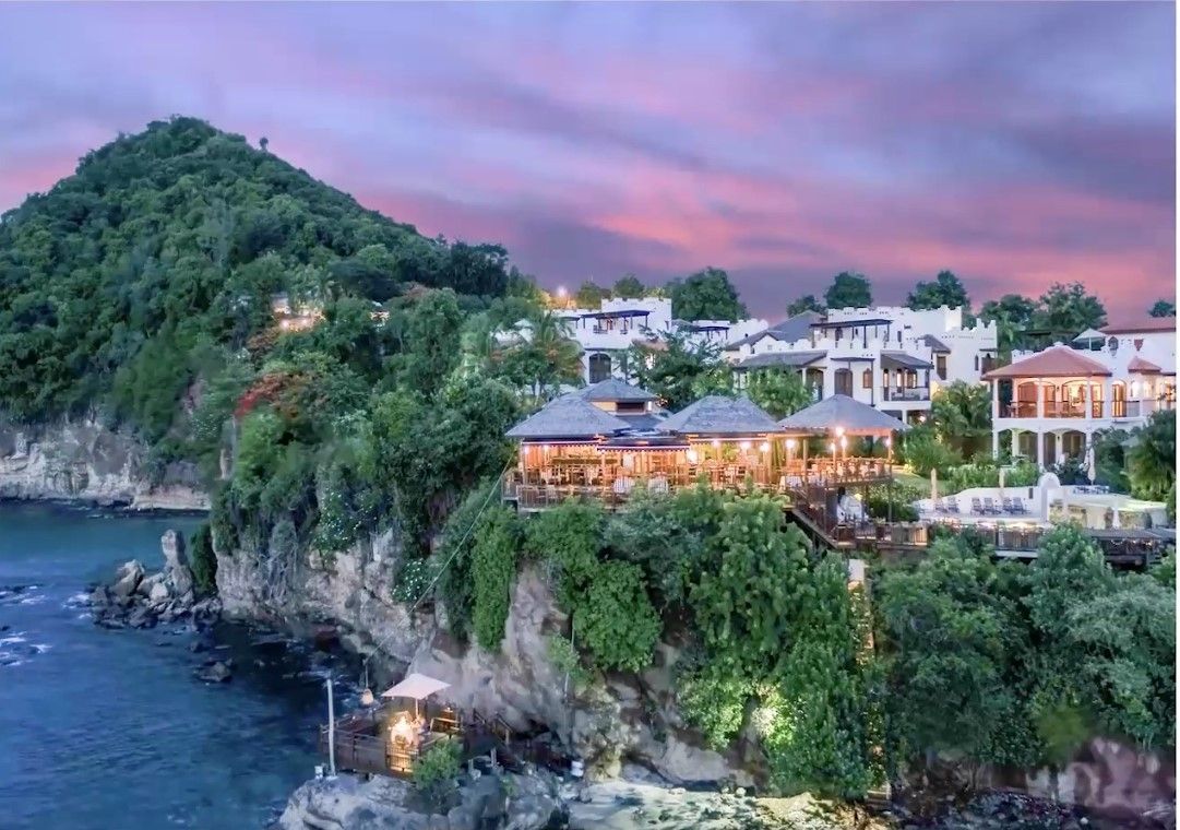 Cliffside seaside resort at dusk, with glowing lights, lush greenery, and pink-purple sky over the ocean