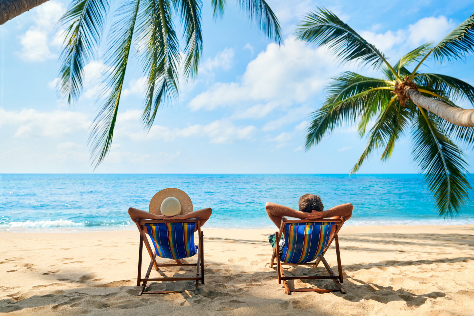 Two people relaxing in beach chairs under palm trees, facing a bright ocean shoreline.