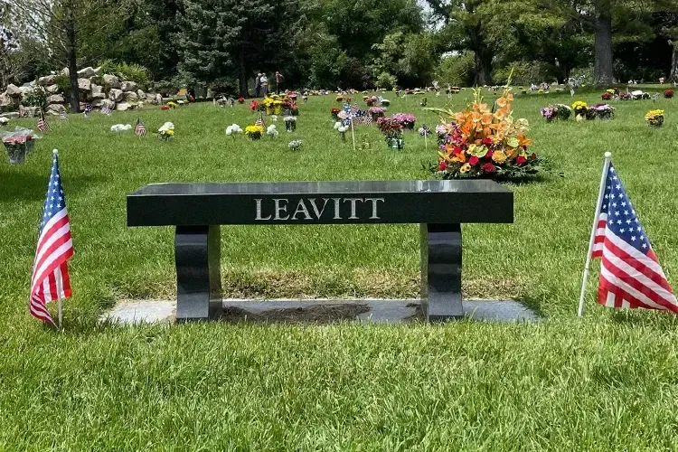 Memorial bench engraved “LEAVITT” with U.S. flags in a grassy garden cemetery.