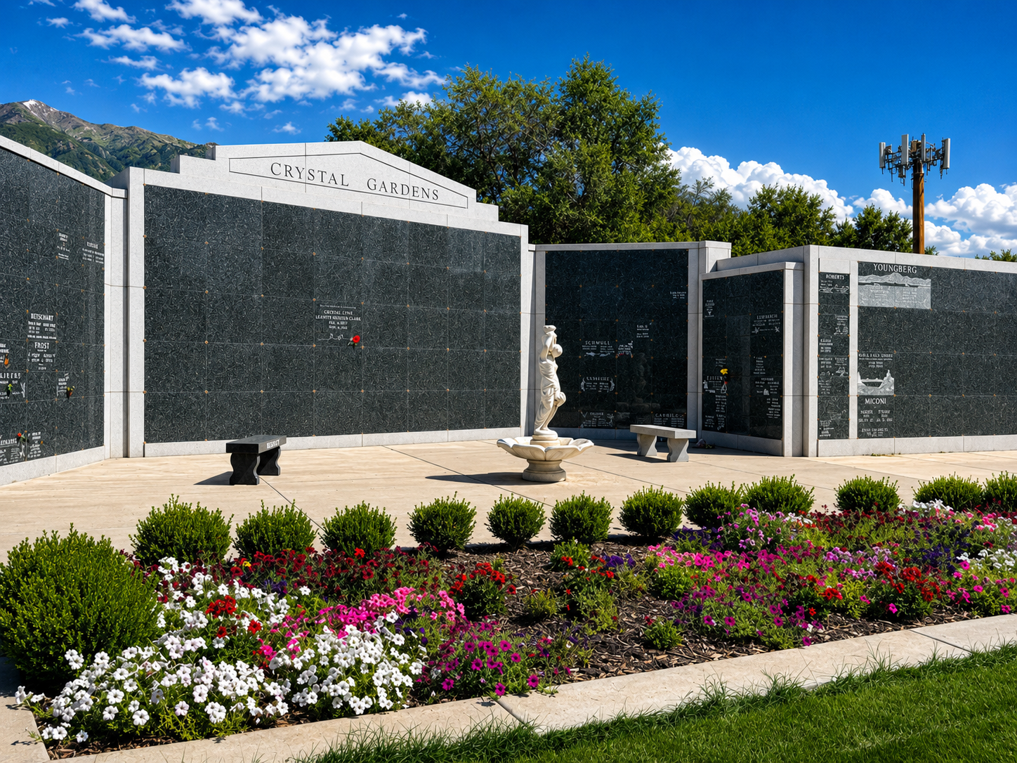 Memorial wall with plaques, white statue, and colorful flower beds under a bright blue sky
