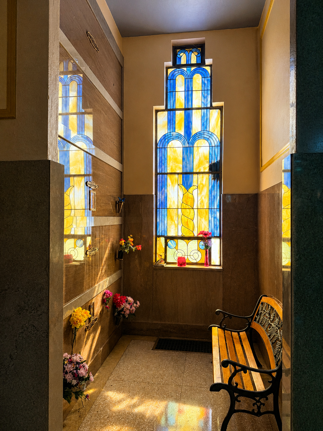 Small chapel alcove with stained-glass window, wooden bench, and flowers along the walls.