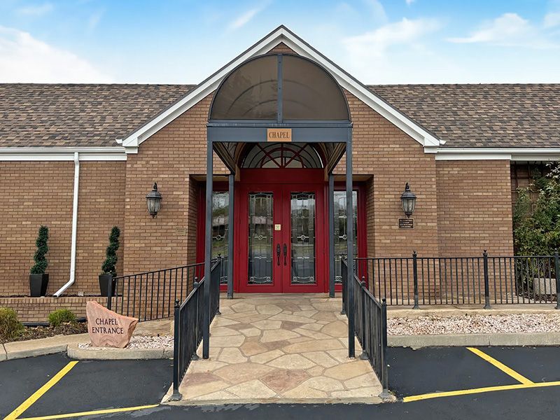 Brick building entrance with red double doors, ramp, and black railings under a peaked awning.